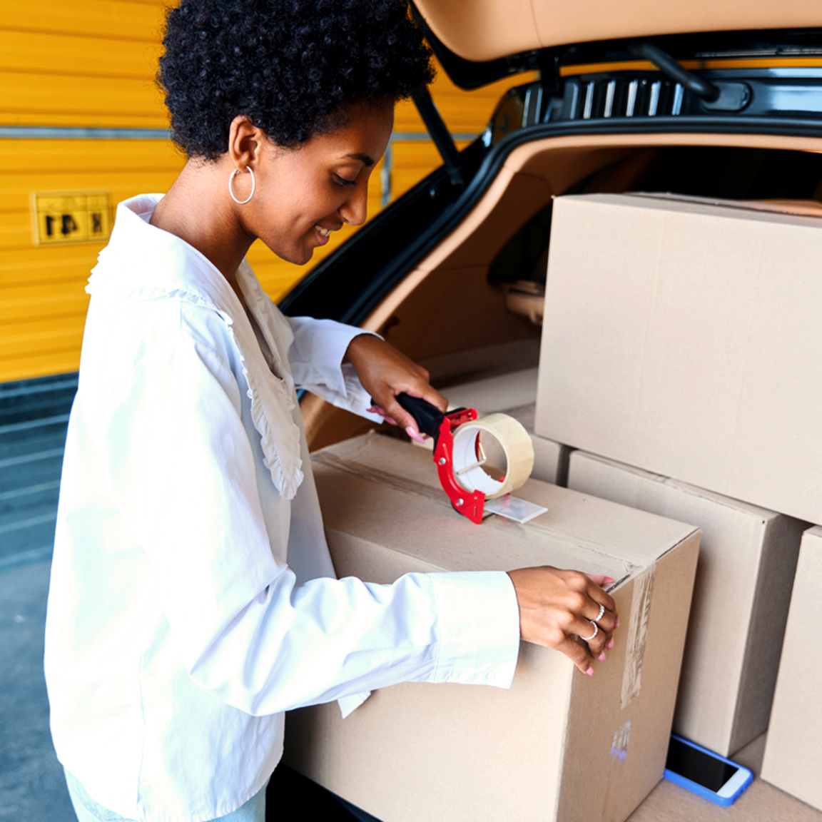 Young woman taping up a box in the back of her car, standing outside of a storage unit.