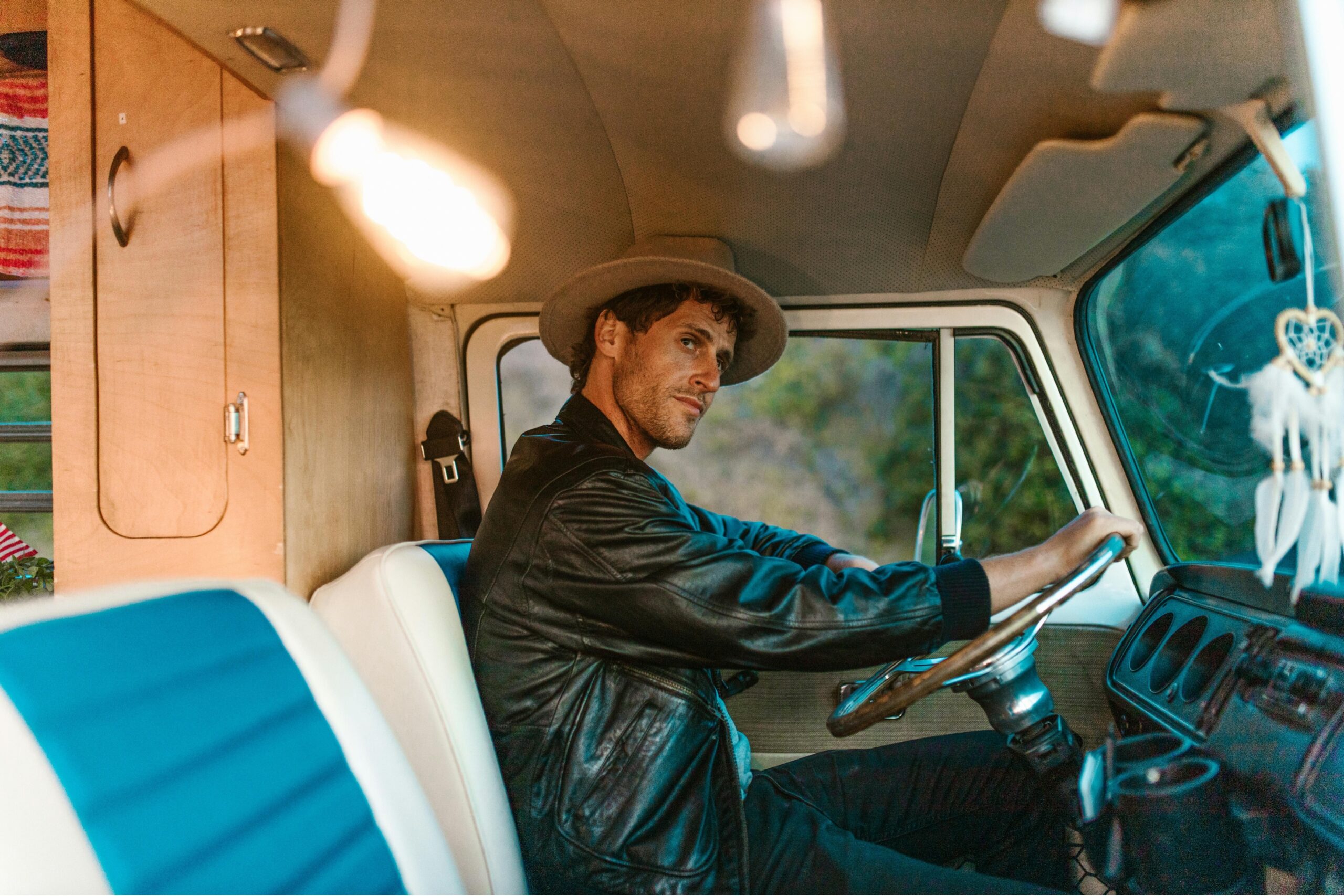 Man in a leather jacket and stylish hat driving an RV with a vintage look.