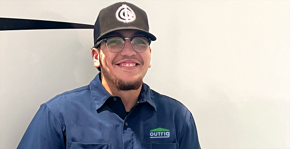 Smiling young man standing in front of an RV wearing a blue shirt, black baseball hat, and glasses.